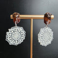 Pendientes de encaje redondo featuring intricate white lace design with wooden and metal accents on gold stand against black background